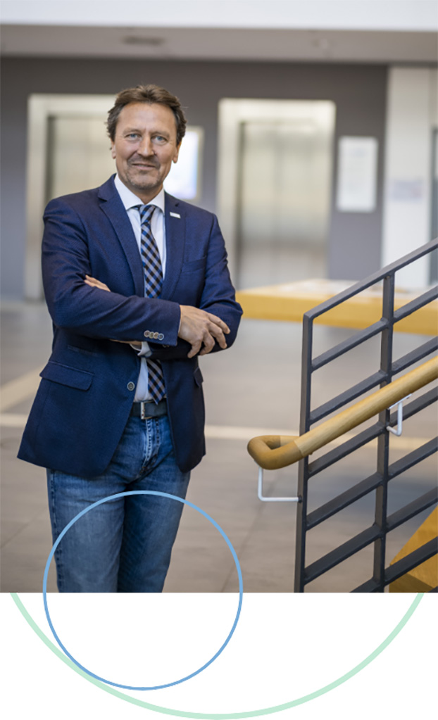 Olaf Guttzeit steht in einem Foyer neben einem Treppenaufgang. Er trägt einen blauen Blazer, weißes Hemd, Krawatte und eine Jeanshose. Seine Arme hält er locker verschränkt.