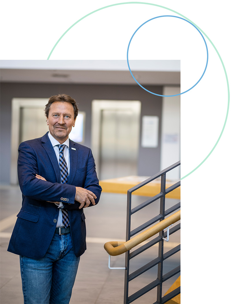 Olaf Guttzeit steht in einem Foyer neben einem Treppenaufgang. Er trägt einen blauen Blazer, weißes Hemd, Krawatte und eine Jeanshose. Seine Arme hält er locker verschränkt.