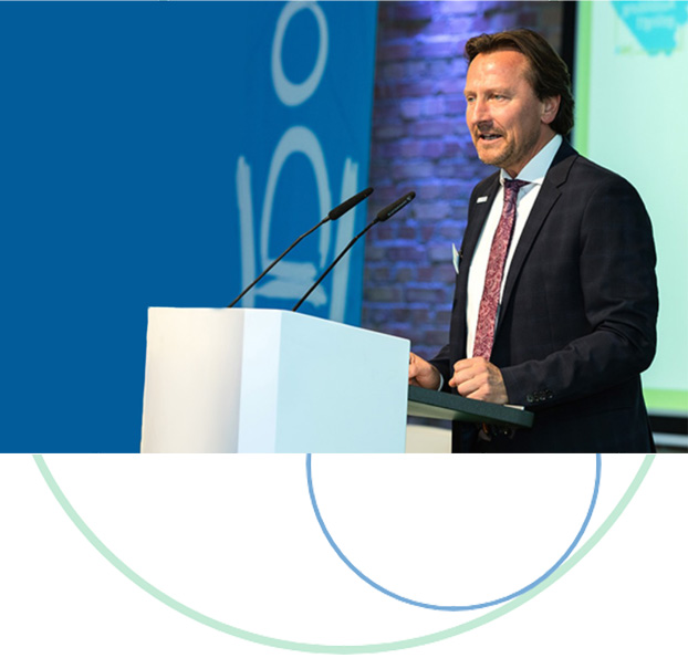 Man sieht Olaf Guttzeit von der Seite. Er trägt einen dunklen Anzug, weißes Hemd und Krawatte. Seine Fäuste liegen leicht geballt auf einem Pult mit zwei Mikrofonen, in die er engagiert spricht.